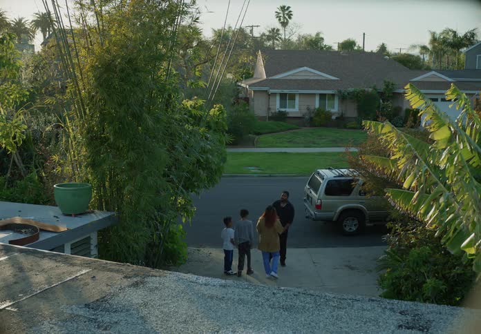 A man and a woman talk to each other while standing outside of a house near a car with two boys nearby