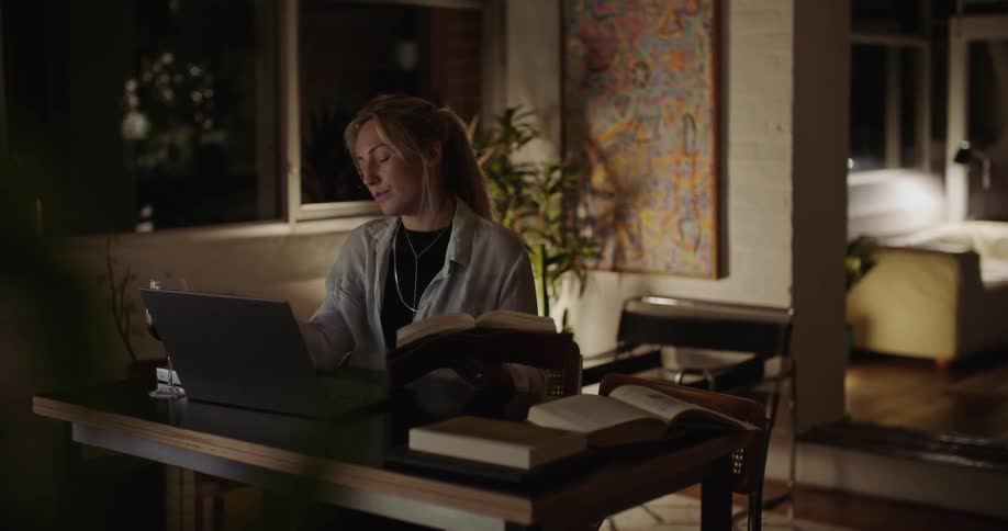 Woman at home drinks wine while reading a book in front of a laptop