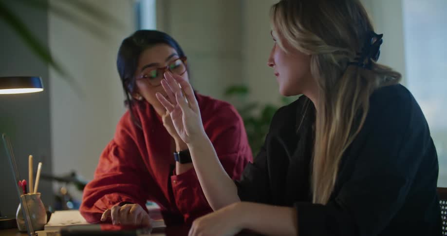 Two women are sitting at a table looking at a laptop computer and pointing to the monitor as they talk