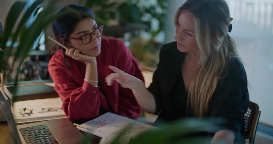 Two women are sitting at the office table watching the laptop screen and talking while working on a project