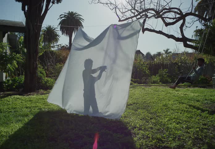 Two boys play in their yard one on a tree swing and one making shadow puppets behind a white sheet