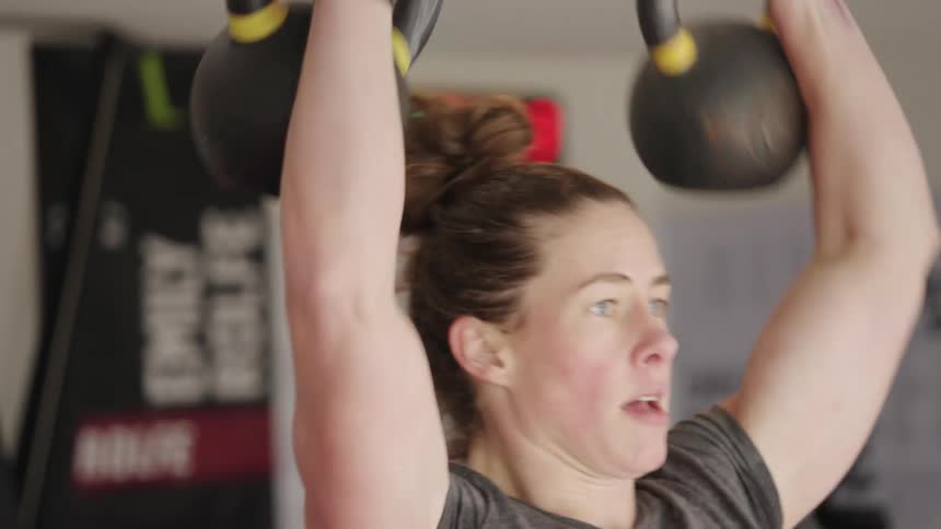 A muscular woman weightlifts kettlebells over her head repeatedly