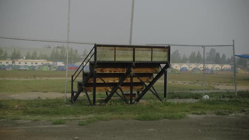 Wide view of old baseball field with old bleachers and many erected tents set up in the background as if for homeless people