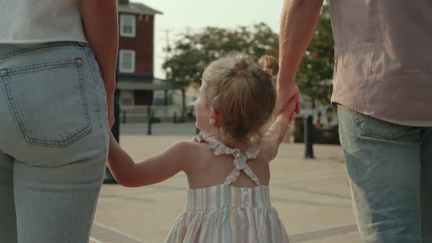 A young girl holds the hands of her mother and father as they walk down an open suburban street