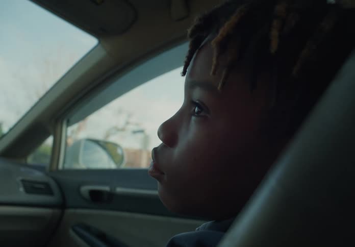 A little boy is sleeping in the passenger seat of an automobile traveling through a neighborhood and he opens his eyes and looks around