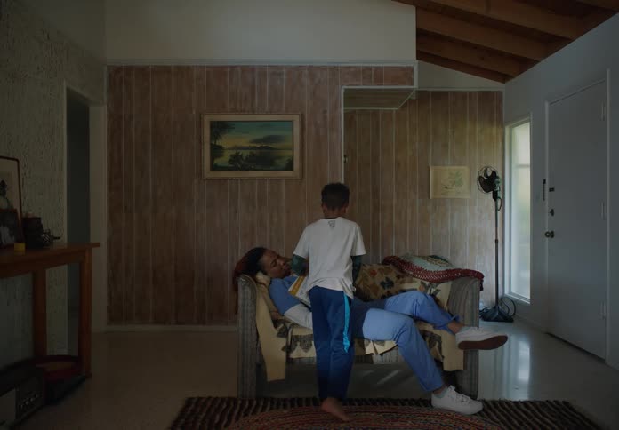 A woman wearing scrubs is sleeping on the couch while her son takes the open book on her chest and returns it to its original place
