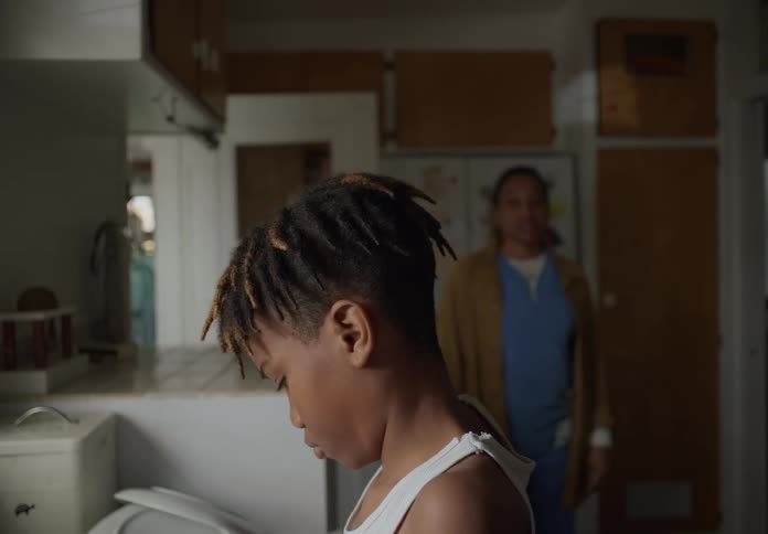 A boy stands in the kitchen near a pile of dishes and a woman enters the room