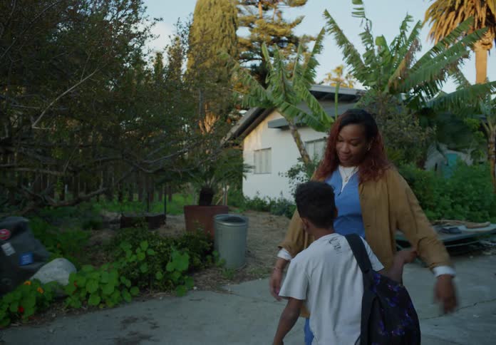 Mother talks to man until her sons join them then she hugs one before they leave with their packed bags