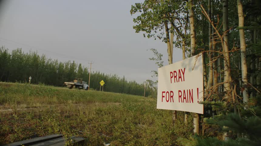 A sign on trees in the woods says to pray for rain as trucks travel on the road