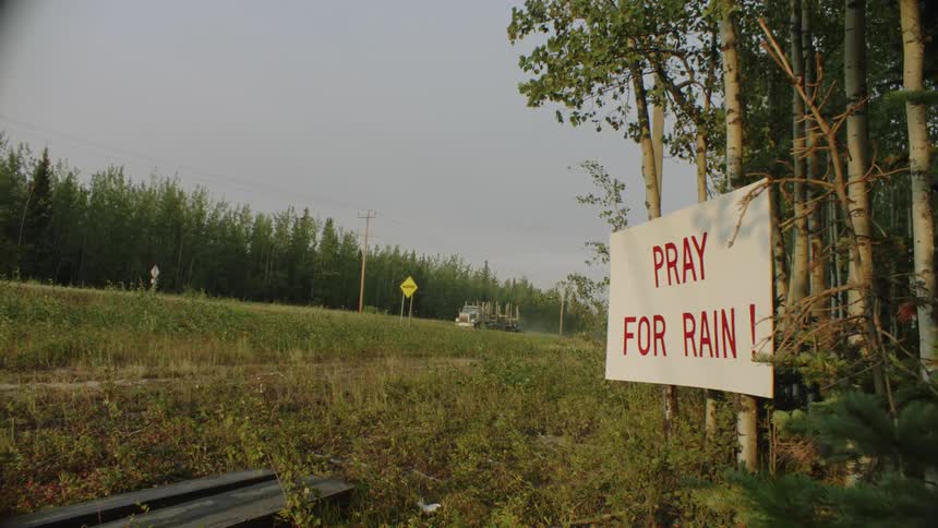 A sign on trees in the woods says to pray for rain as a truck travels on the road
