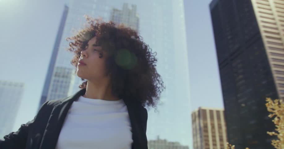 Young woman standing against backdrop of skyscrapers during the day with sun flare coming in between buildings