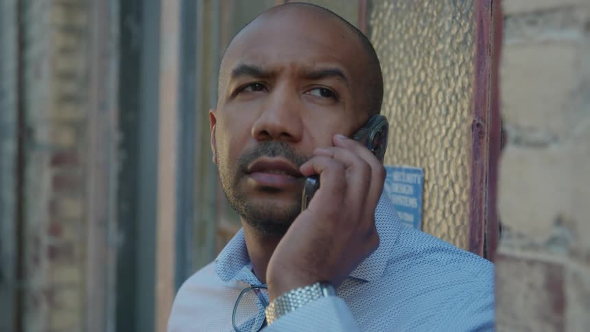 A man standing up against a brick building and windows talks on a cell phone