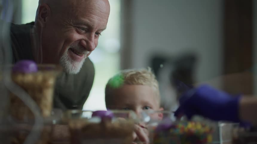 A grandfather and grandson in a shop happily order candy scooped out of a bowl by a gloved hand