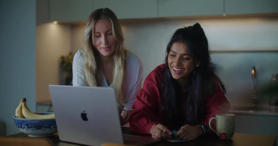 Two young women at a kitchen table laugh at something on a laptop and take notes on paper