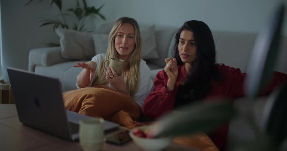 Two friends have a conversation while sitting on the living room floor watching a show on the laptop at home