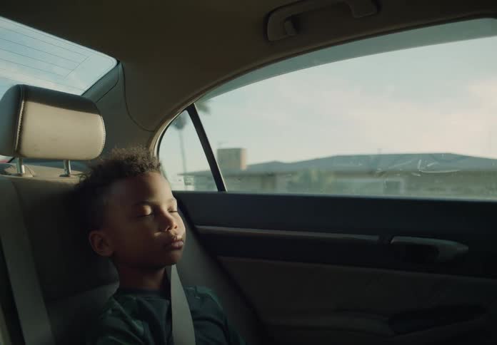 Close up of a young boy wearing a seatbelt asleep in the backseat of a car driving at morning