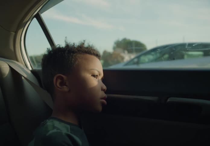 A little boy sitting in the back of a car as it is driving the sun on his face as he looks sad