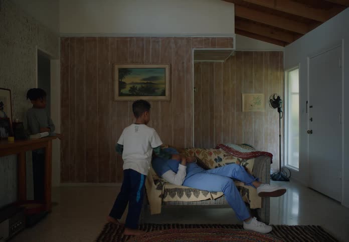 A boy walks in the house and puts a coat over his mom sleeping on the couch