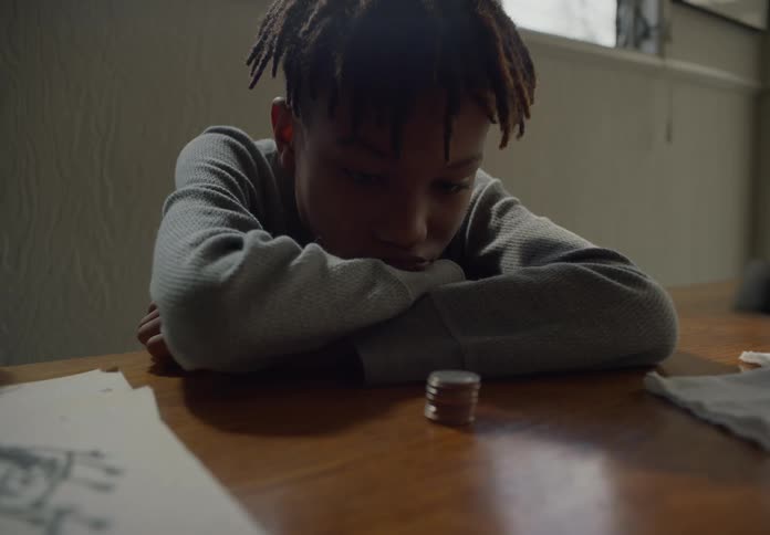 A boy sits at the table with another boy and stares at a stack of coins then knocks them over and spreads them out