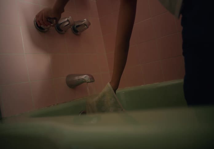 A shower and tub as a young boy turns it on and lets water run on a towel in his hand before he walks away