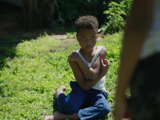 A young boy sits on the grass holding his arms close while glaring