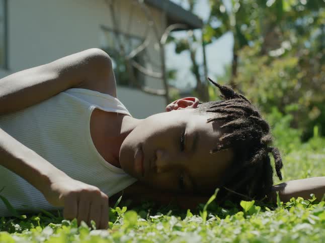 Boy with dreadlocks and white undershirt lies on green lawn