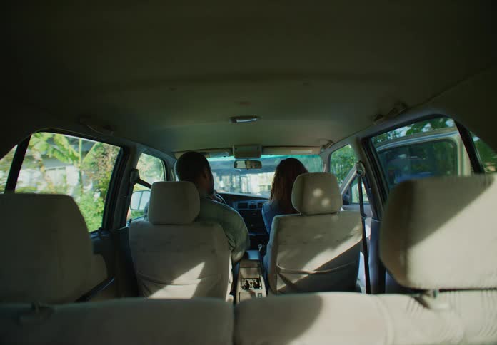 A man is sitting in the drivers seat of an automobile as his wife is walking down their driveway and she gets in the car