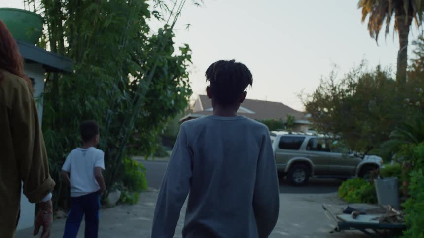 A mother and her two children walk through a garage full of furniture out to the driveway to meet their father who is walking from his truck towards them