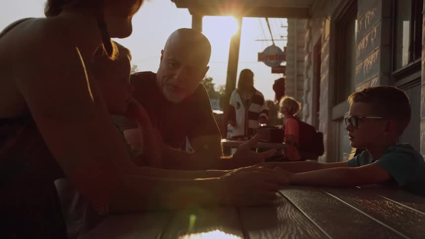 A young boy sits at a table across from his father brother and mother outside of a restaurant