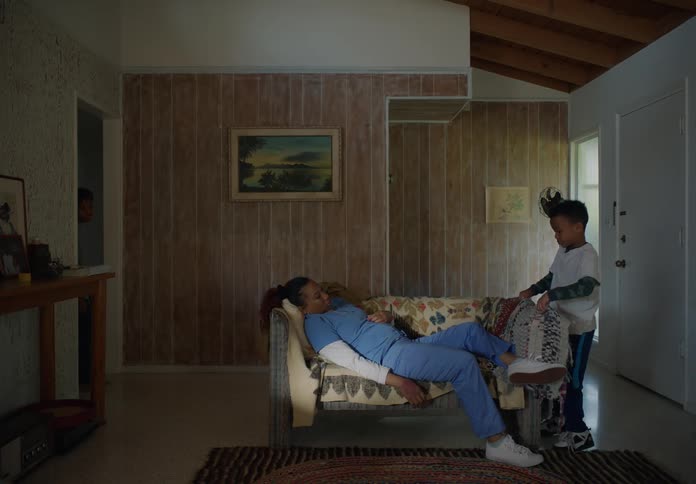A boy takes a book from his mother and covers her with a blanket as she dozes on the couch