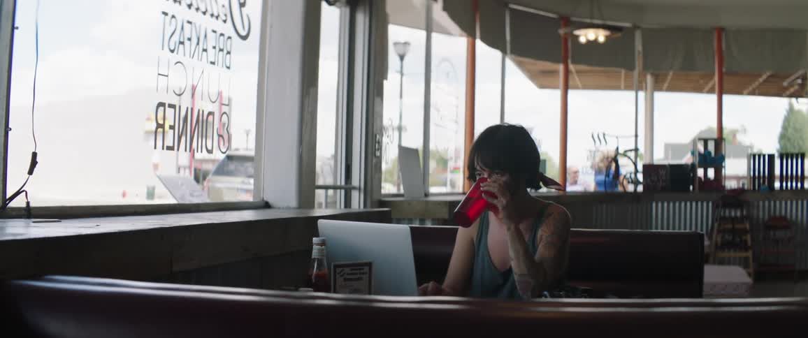 A woman sits at a table in a diner drinking water from a red cup