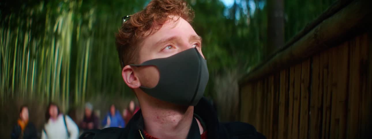 A young man wearing a medical mask takes a photograph with a camera while standing on a path through a bamboo forest during the day