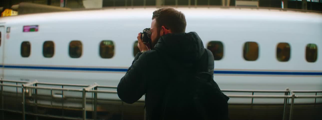 Male photographer with a beard a black coat and backpack takes photographs of a train arriving at the station
