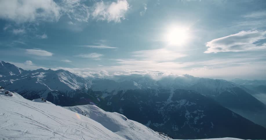 The sun shines brightly over snow covered mountains under a cloudy sky