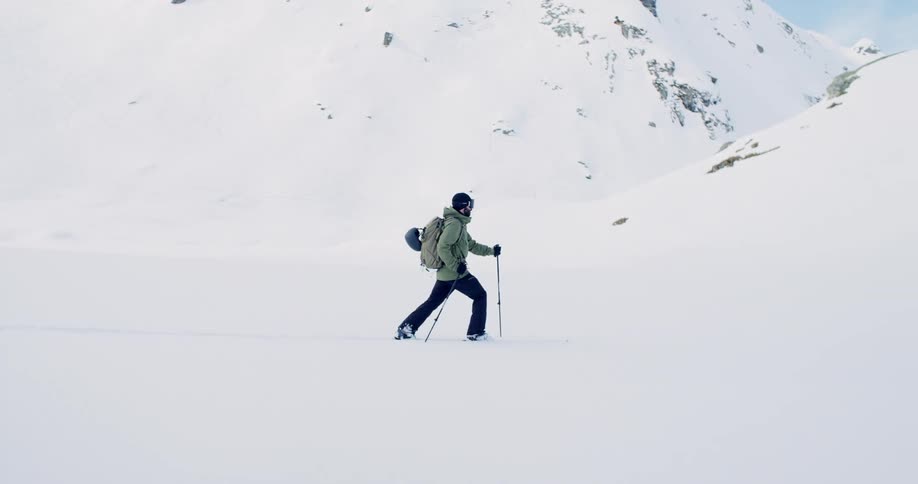 A mountaineer uses trekking poles to walk through a snowy field in the mountains during the day