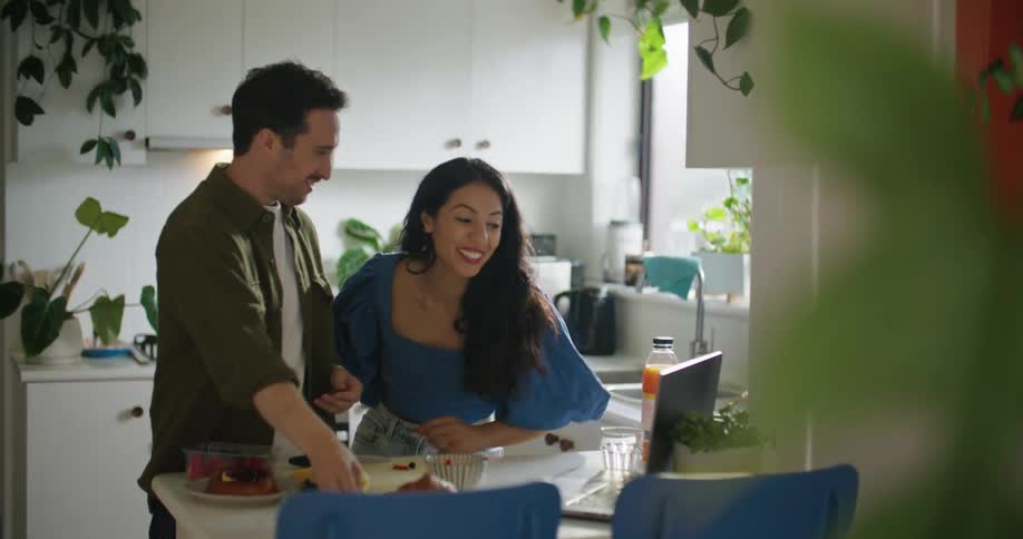 Young couple eating breakfast in the kitchen at home greeting someone on a video call