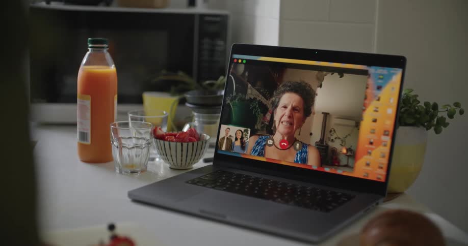 An elderly woman is on a laptop having a conversation with a person almost completely off camera in a kitchen