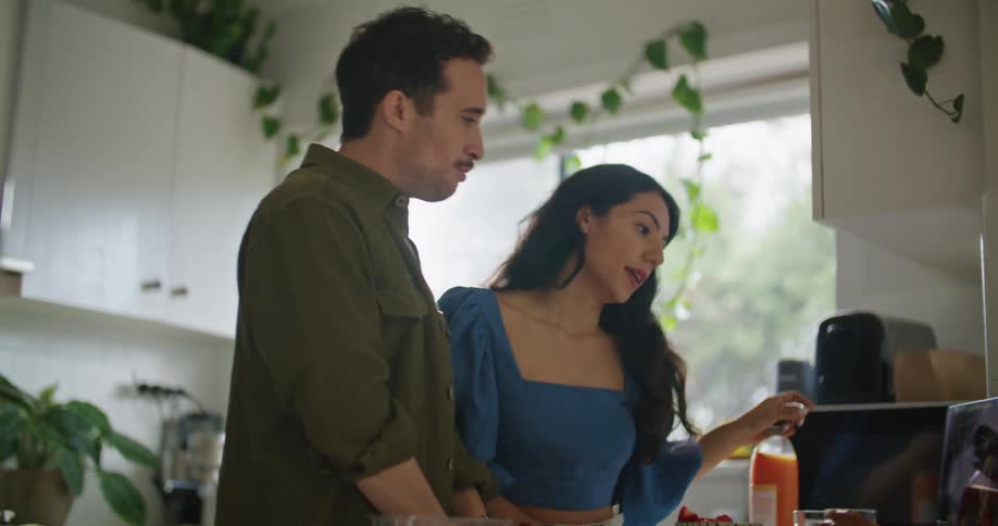 Man and woman standing in the kitchen discussing something on their laptops and having breakfast