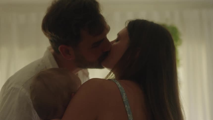 A mother and father kissing each other while holding a baby in their living room