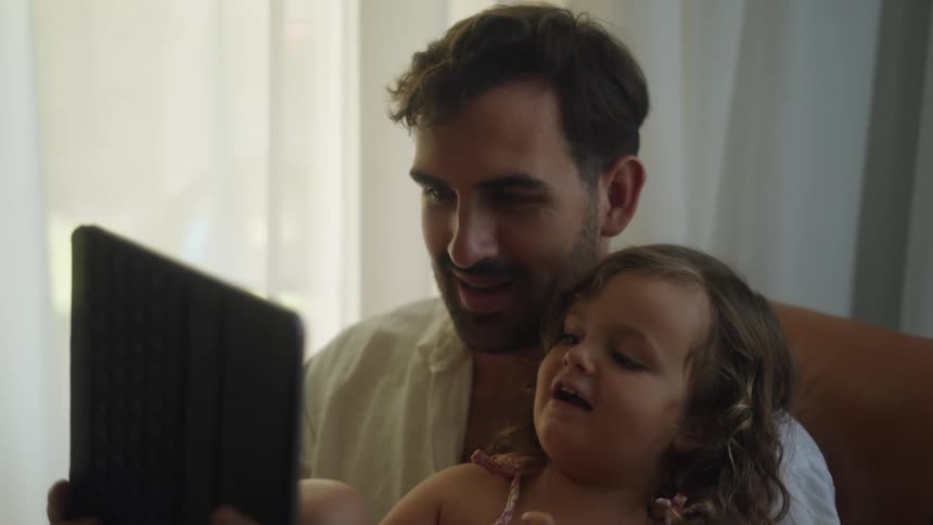 A toddler sits on her dads lap as he holds a tablet while she writes on it with a stylus