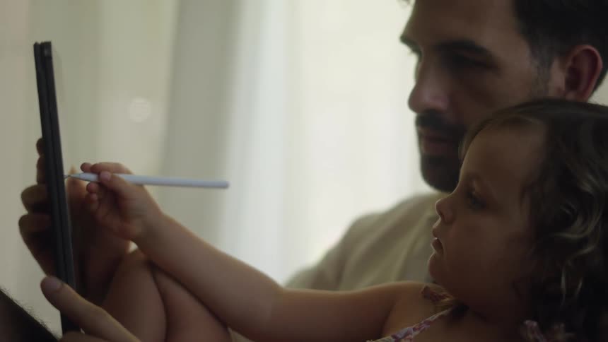 A young girl uses a stylus on a computer tablet while being held in the arms of a man sitting indoors during the day