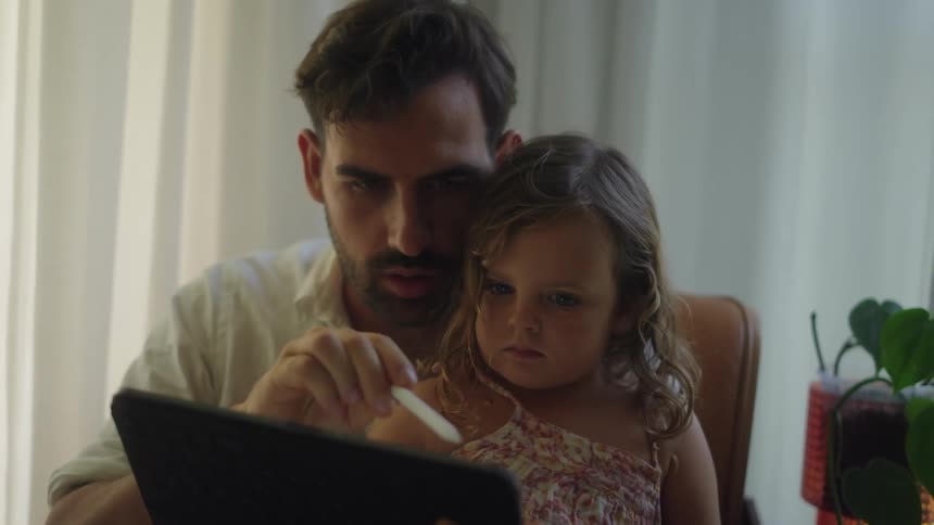 A man holds his daughter in his lap while using a tablet
