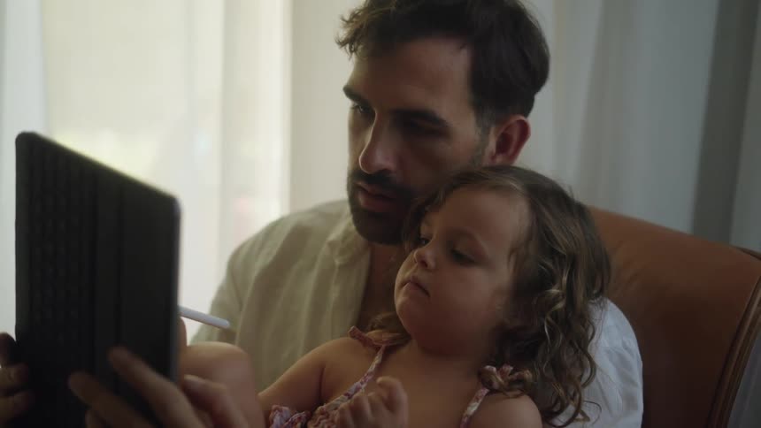 A man sitting indoors holds a young girl in his arms while the girl uses a stylus to operate a computer tablet