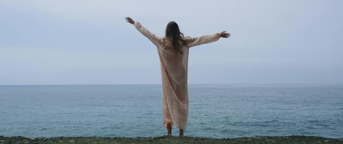 A woman standing on a rocky shore oceanside raises her hands above her head in a stretch