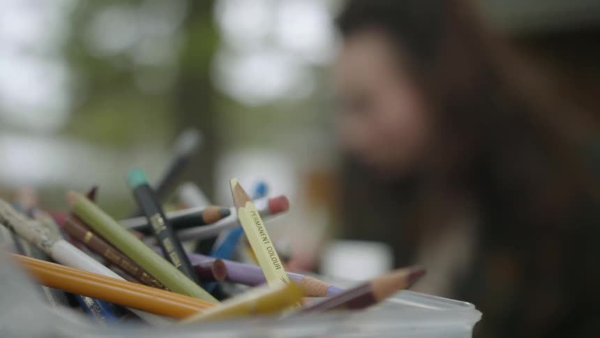 Close up of a plastic container full of color pencil next to a woman drawing outdoors
