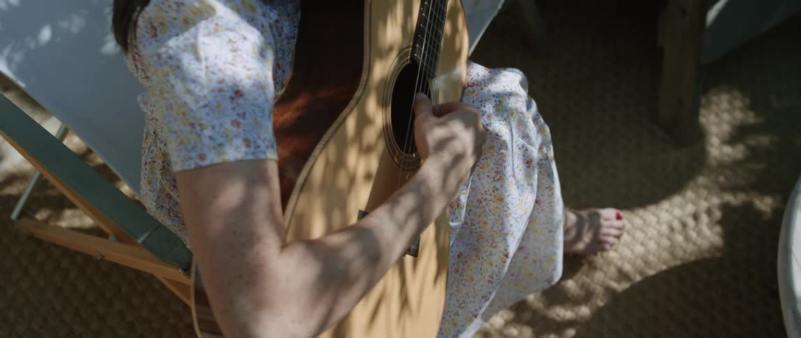 The sun shines upon a woman wearing a dress sitting outside playing an acoustic guitar