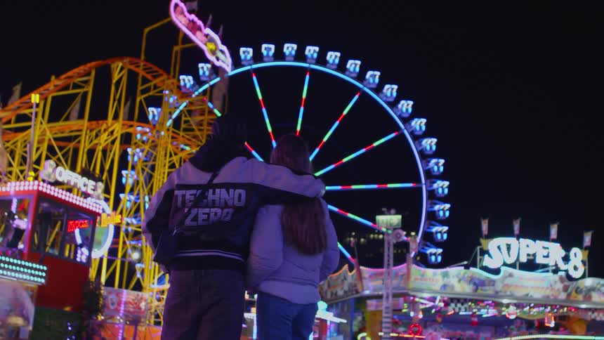 A couple is looking up at a ferris wheel with bright lights at a carnival