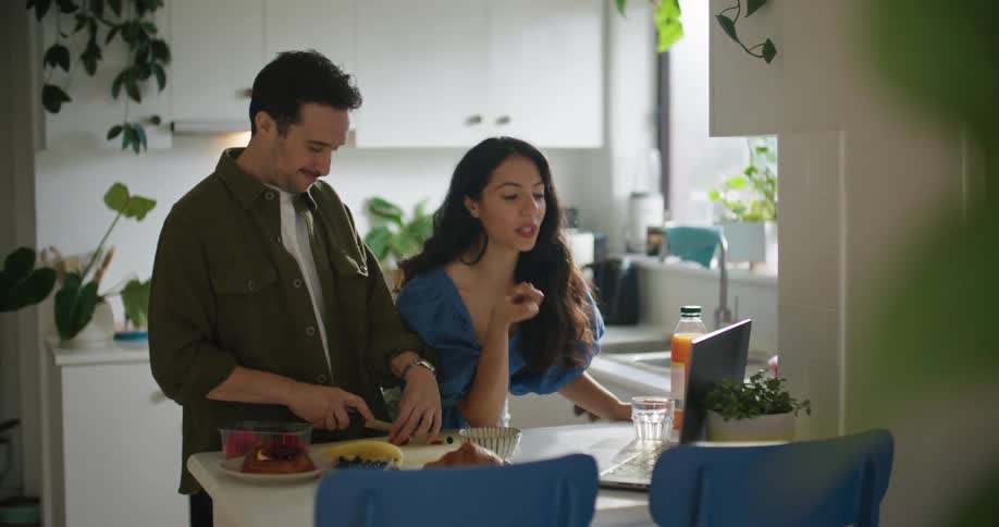 A man and a woman at a kitchen table talk to someone through a laptop while cutting food with a knife