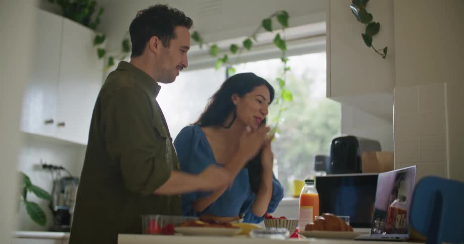 A man and a woman at a kitchen table with food and drinks talk to someone through a laptop