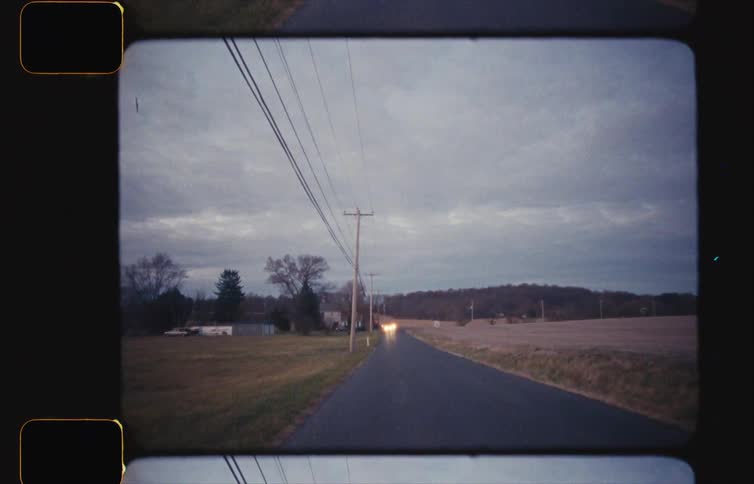 A narrow road passes by a wide open grass field as a few cars are in the distance